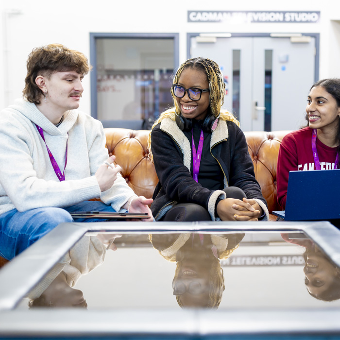 A male student is chatting to two female students. They are smiling and sitting on a brown sofa in an open studio space