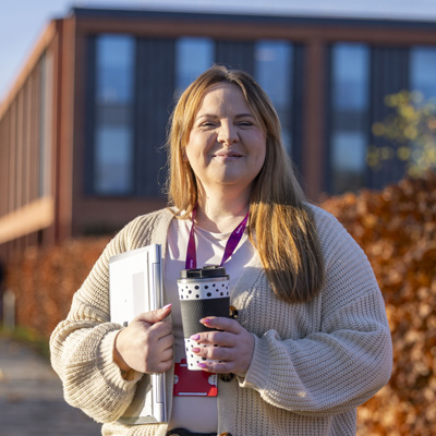 A female with long blonde hair is holding a notebook and coffee outside