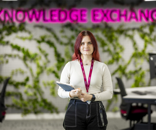 A female student with long red hair is standing holding a notebook and pen