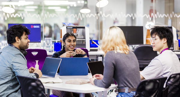 A group of diverse students are sitting at a table with their laptops chatting and smiling in the Smart Zone