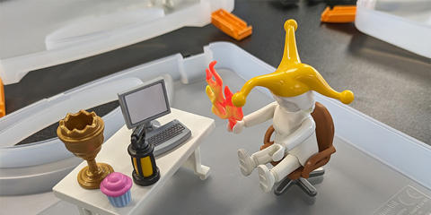 A Playmobil figure sat at a desk holding flames and wearing a jester's hat