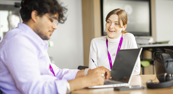 A young female dressed smartly is sitting at a desk, collaborating with a young male using a laptop